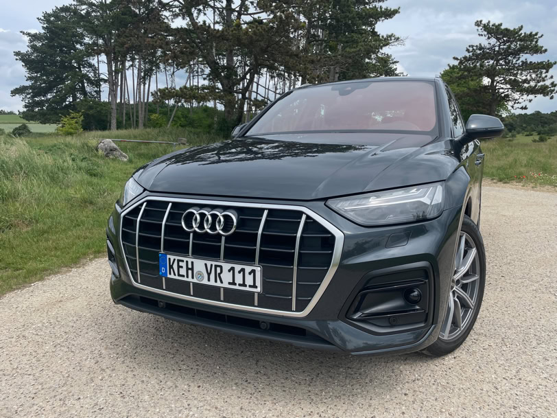 Ein schwarzer Audi SUV steht auf einem Schotterweg, im Hintergrund sind Bäume und ein bewölkter Himmel zu sehen.